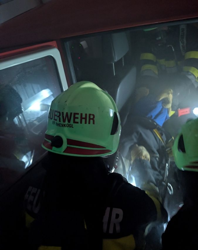 Ein Feuerwehrmann mit einem grünen Helm, auf dem 'Feuerwehr' steht, ist in einem Fahrzeug, möglicherweise einem Feuerwehrwagen, mit anderen Feuerwehrleuten um ihn herum.