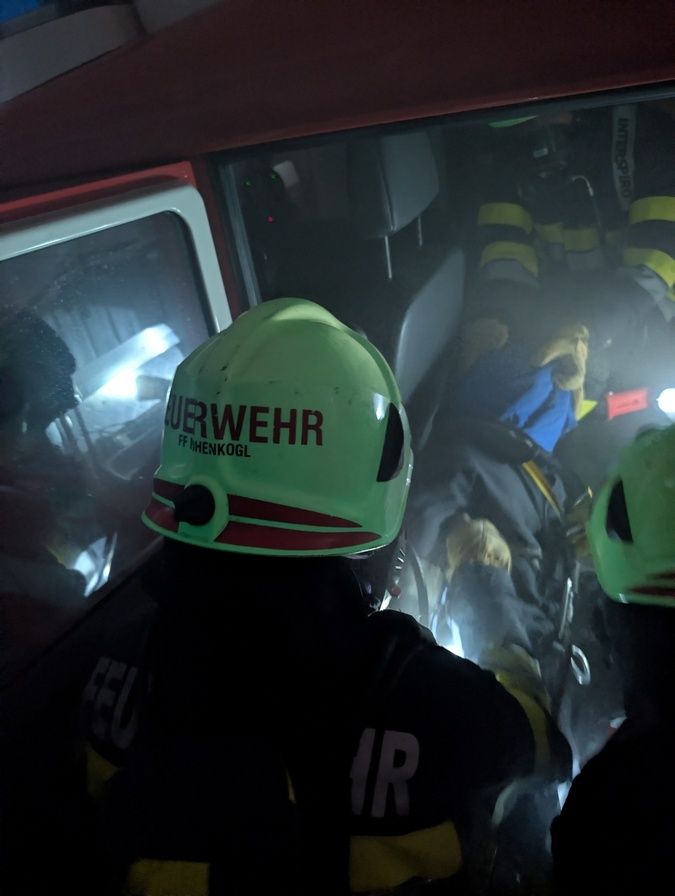Ein Feuerwehrmann mit einem grünen Helm, auf dem 'Feuerwehr' steht, ist in einem Fahrzeug, möglicherweise einem Feuerwehrwagen, mit anderen Feuerwehrleuten um ihn herum.