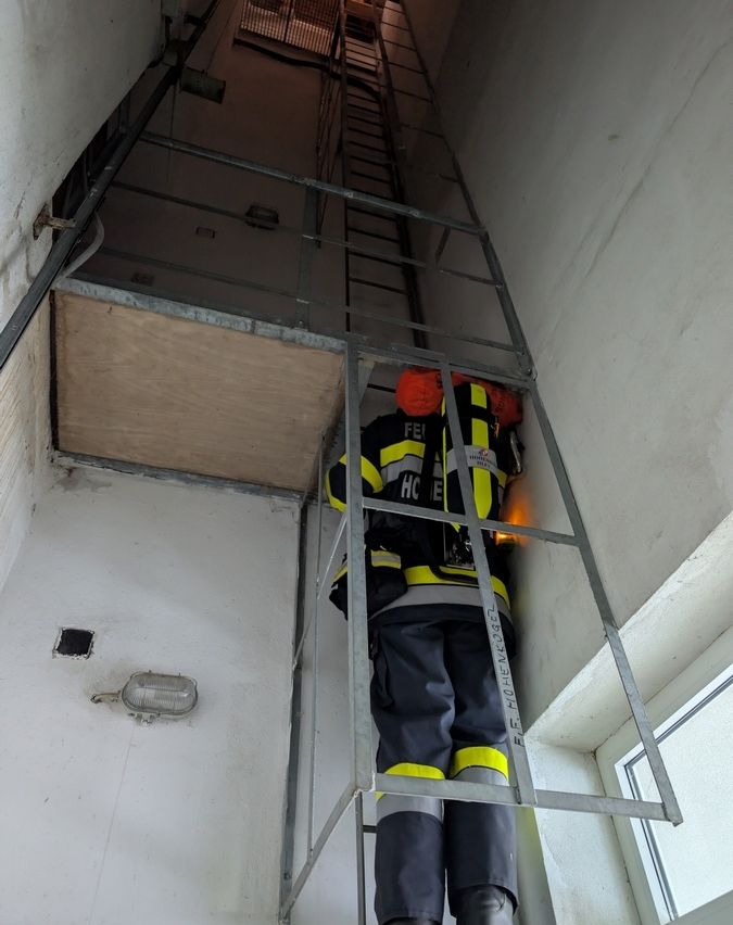 Ein Feuerwehrmann in voller Ausrüstung klettert eine Metallleiter in einem Gebäude. Die Leiter ist an einer Wand mit einem Holzpaneel und einer Lichtarmature befestigt.