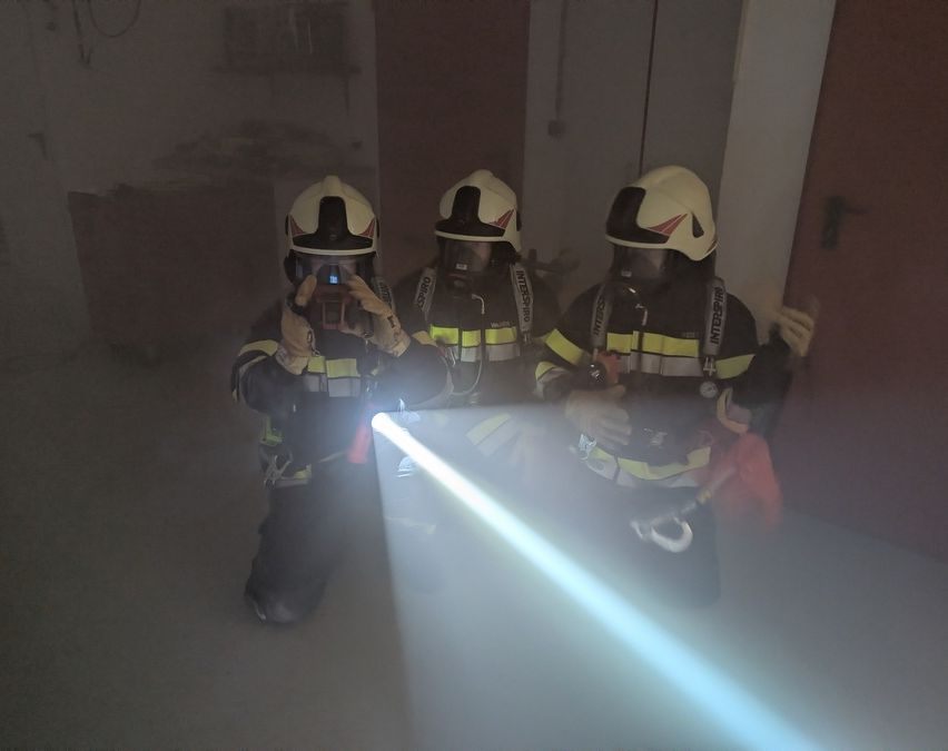Drei Feuerwehrleute in Schutzkleidung werden von einer Taschenlampe beleuchtet und stehen in einem rauchigen Raum mit einer Tür im Hintergrund.