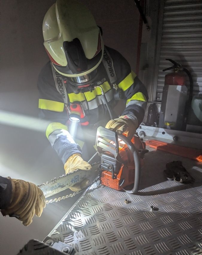 Ein Feuerwehrmann mit Helm und Handschuhen benutzt eine Kettensäge auf einer Metalloberfläche.