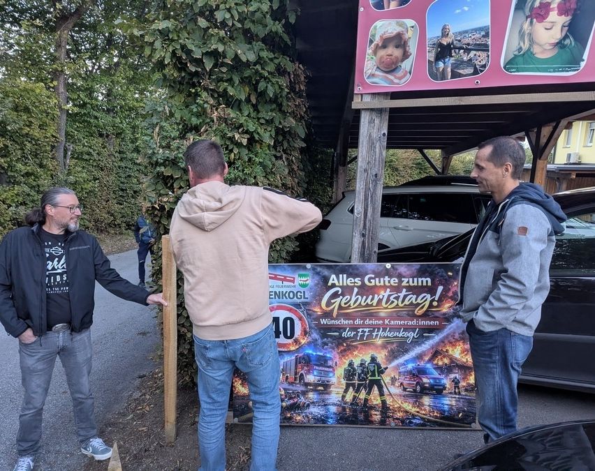 Drei Männer stehen vor einem Banner mit einem Feuerwehrauto-Bild, einer zeigt darauf. Dahinter ist ein Auto unter einem Vordach geparkt.