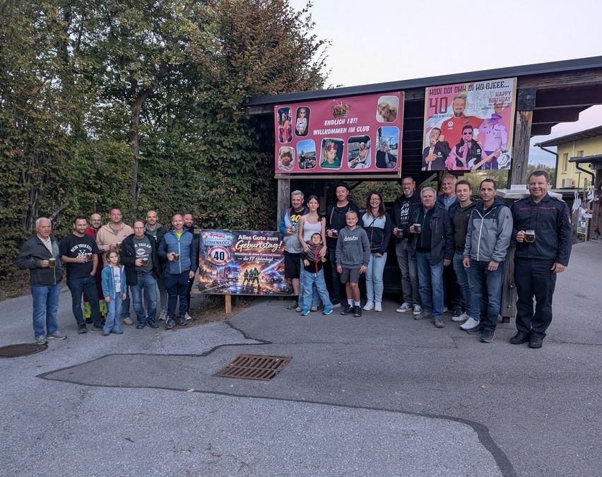 Eine Gruppe von Menschen steht vor einem dekorierten Stand mit Bannern und Plakaten und feiert ein Ereignis.