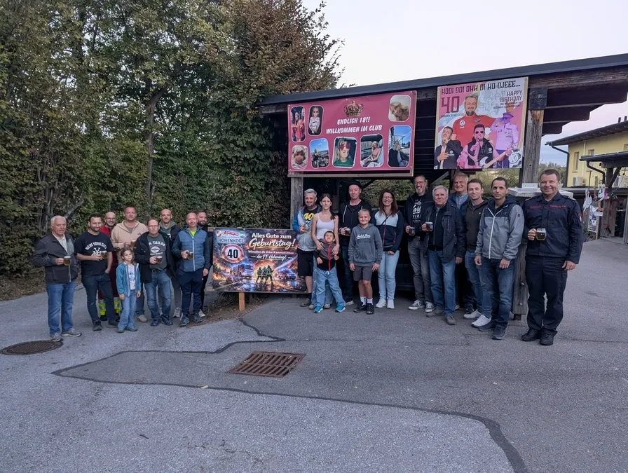 Eine Gruppe von Menschen steht vor einem dekorierten Stand mit Bannern und Plakaten und feiert ein Ereignis.