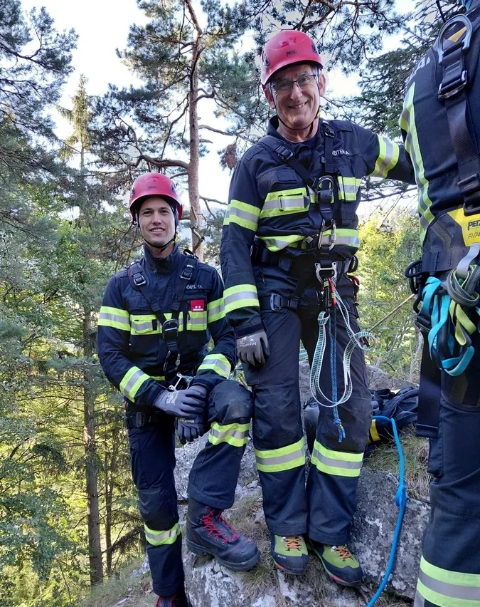 Drei Feuerwehrleute in voller Ausrüstung, zwei stehen auf einem Felsen und einer lehnt sich an einen Baum, lächeln für ein Foto im Wald.
