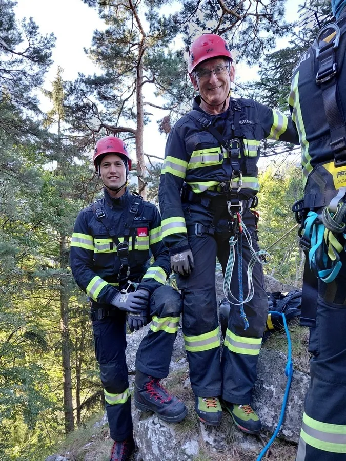 Drei Feuerwehrleute in voller Ausrüstung, zwei stehen auf einem Felsen und einer lehnt sich an einen Baum, lächeln für ein Foto im Wald.