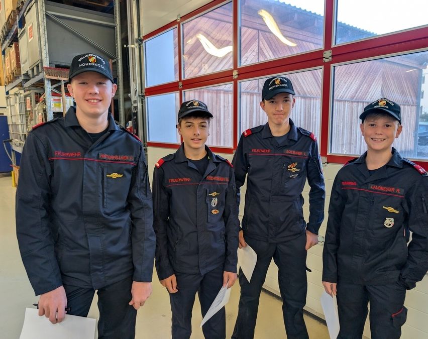 Vier junge Männer in dunkelblauen Uniformen mit rotem Rand und schwarzen Mützen mit weißen Logos stehen in einer Reihe vor einem Gebäude.