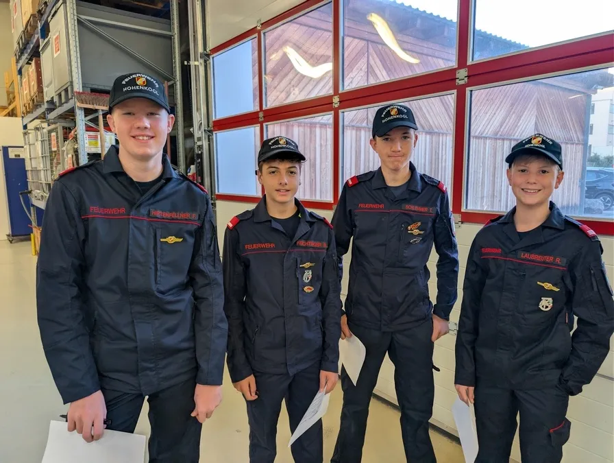 Vier junge Männer in dunkelblauen Uniformen mit rotem Rand und schwarzen Mützen mit weißen Logos stehen in einer Reihe vor einem Gebäude.