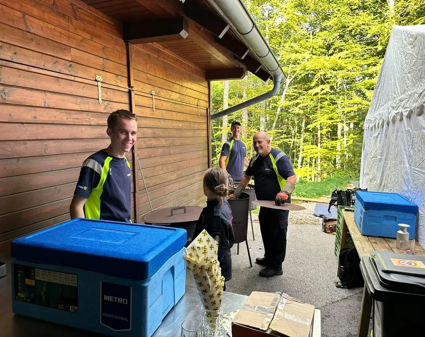 Drei Männer und ein junges Mädchen stehen vor einem hölzernen Gebäude. Die Männer tragen Uniformen und scheinen zu arbeiten. Das Mädchen schaut auf etwas am Boden.