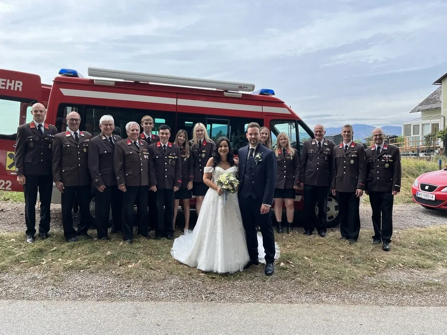 Eine Hochzeitsgesellschaft steht vor einem roten Feuerwehrauto mit der Nummer 22. Die Braut und der Bräutigam stehen in der Mitte, umgeben von uniformierten Offizieren.