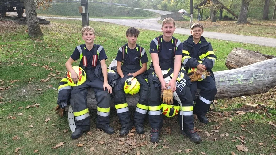 Vier junge Männer in Feuerwehruniformen sitzen auf einem Baumstamm in einem Park.