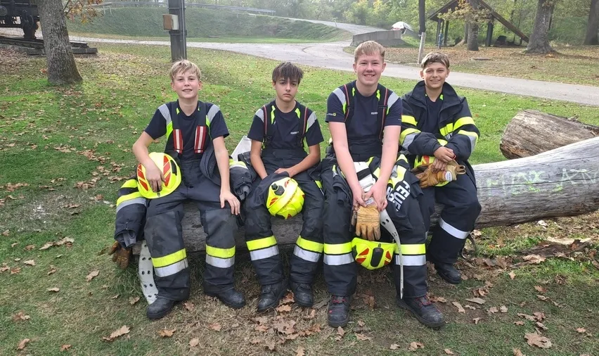 Vier junge Männer in Feuerwehruniformen sitzen auf einem Baumstamm in einem Park.