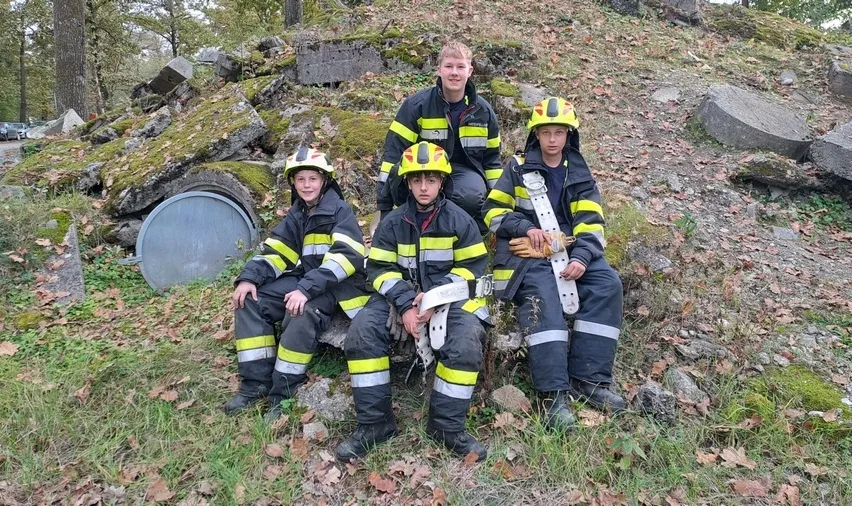 Fünf junge Feuerwehrleute sitzen auf einem Felsen im Wald, in gelben und schwarzen Uniformen mit Helmen.
