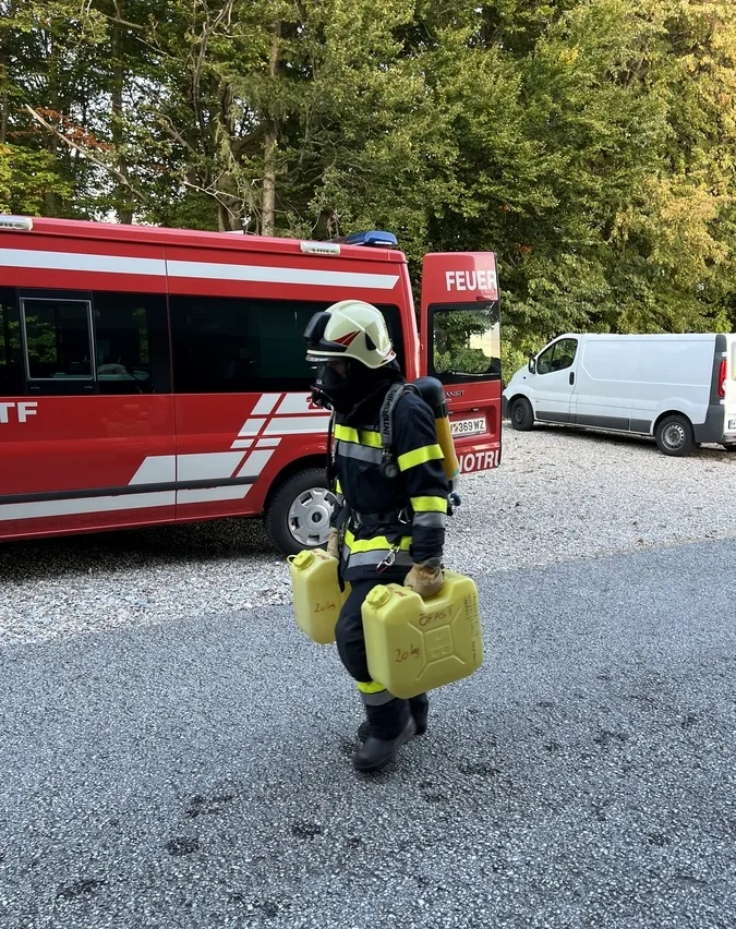 Ein Feuerwehrmann geht auf einem Kiesweg und trägt zwei gelbe Kanister. Dahinter steht ein roter Feuerwehrwagen mit dem Wort 'Feuer' darauf. Rechts davon ist ein weißer Lieferwagen geparkt.