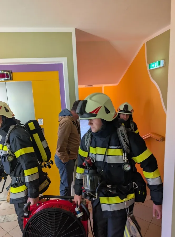 Eine Gruppe von Feuerwehrleuten in voller Ausrüstung, einschließlich Helmen, geht durch einen Flur. Einer von ihnen trägt einen rot-schwarzen Ventilator.