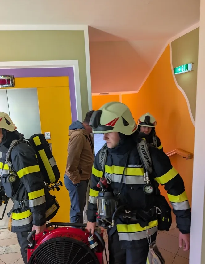 Eine Gruppe von Feuerwehrleuten in voller Ausrüstung, einschließlich Helmen, geht durch einen Flur. Einer von ihnen trägt einen rot-schwarzen Ventilator.