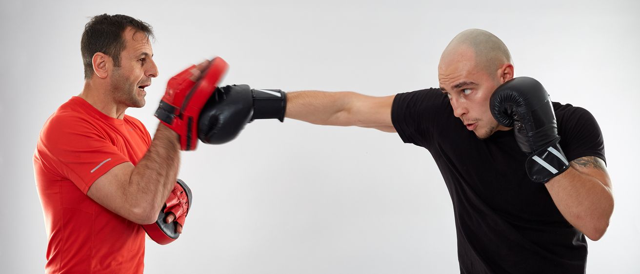 Zwei Männer in einem Studio, einer in Rot und einer in Schwarz, führen einen Boxkampf. Der Mann in Rot trägt Handschuhe und wirft einen Schlag.