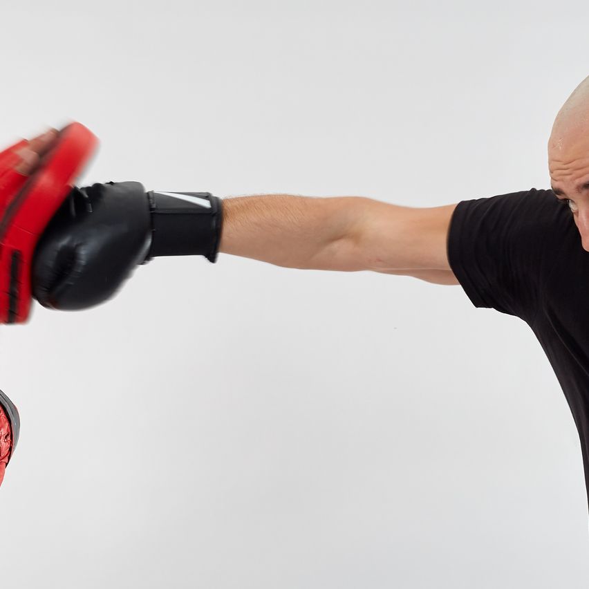 Zwei Männer in einem Studio, einer in Rot und einer in Schwarz, führen einen Boxkampf. Der Mann in Rot trägt Handschuhe und wirft einen Schlag.