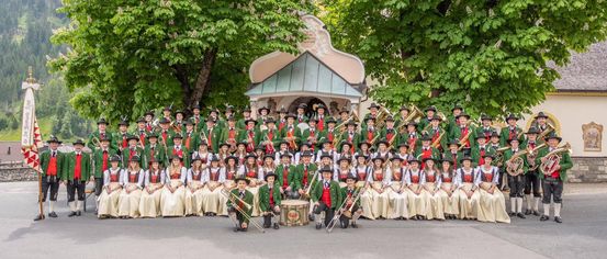 Eine große Gruppe von Musikern in traditioneller Kleidung posiert für ein Foto vor einer kleinen Kapelle mit Bäumen im Hintergrund.
