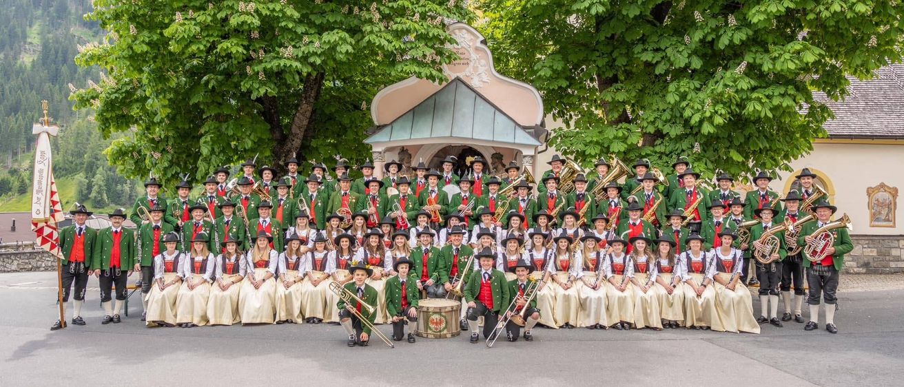 Eine große Gruppe von Musikern in traditioneller Kleidung posiert für ein Foto vor einer kleinen Kapelle mit Bäumen im Hintergrund.