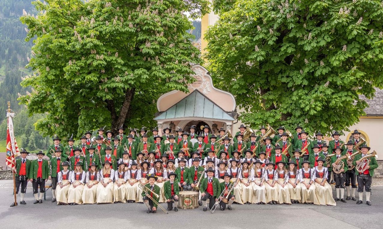 Eine große Gruppe von Musikern in traditioneller grüner und roter Kleidung, die vor einem Pavillon mit Bäumen im Hintergrund für ein Foto posieren.