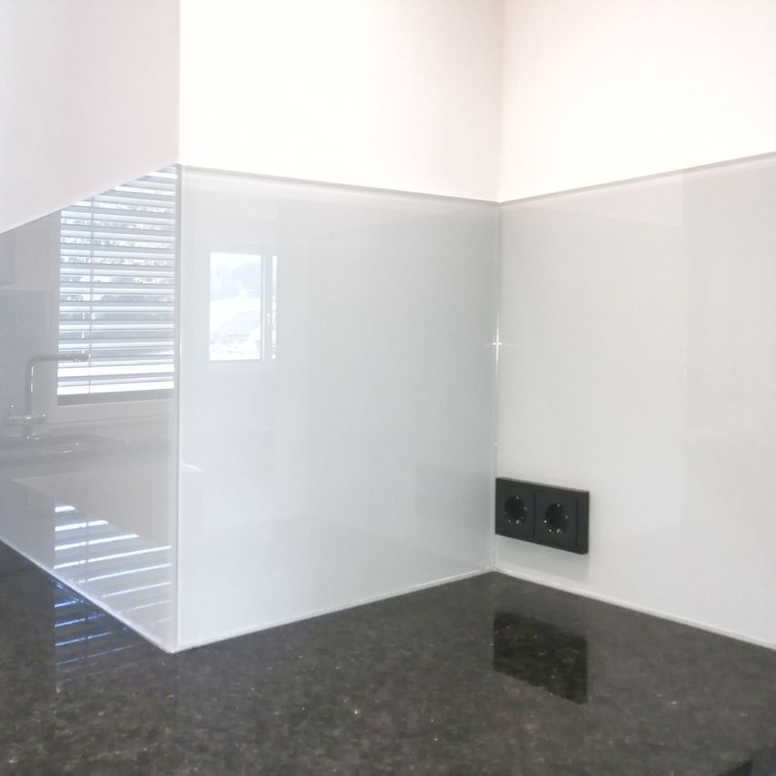 Eine moderne Küche mit weißen Glasplatten, einer Granitarbeitsplatte und einem Fenster mit Jalousien. An der Wand ist eine Steckdose montiert.