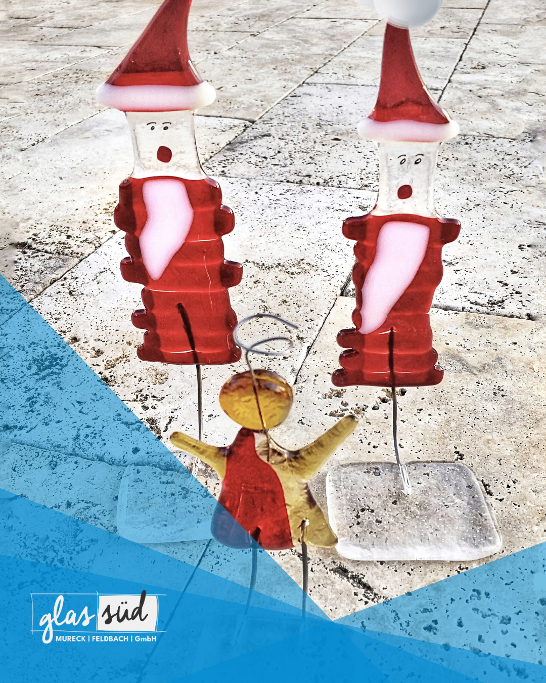 Zwei rote Glasfiguren von Santa Claus und einem Kind stehen auf einer Betonoberfläche. Das Kind trägt ein rotes Kleid.