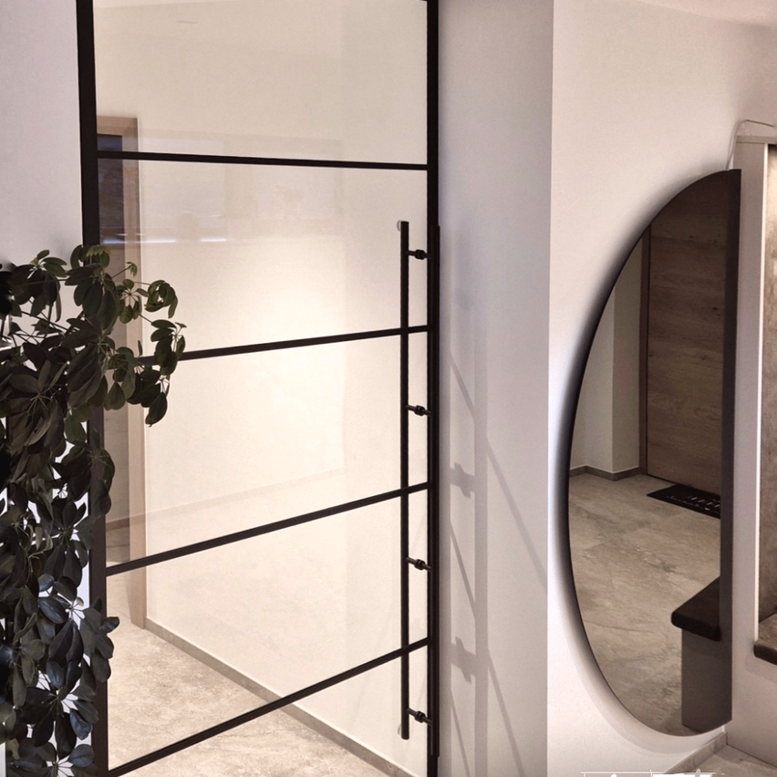 Ein Innenraum zeigt eine moderne Glastür mit schwarzem Rahmen und einem reflektierenden Spiegel an einer weißen Wand.