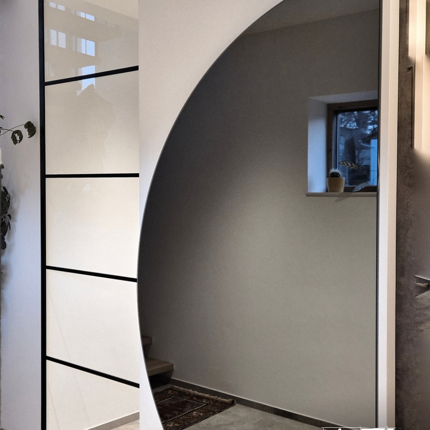 Ein moderner Raum mit einer geschwungenen Glastür, einer weißen Wand und einem Fenster mit einem Topf auf der Fensterbank. Ein kleiner Teppich liegt in der Nähe der Treppe.