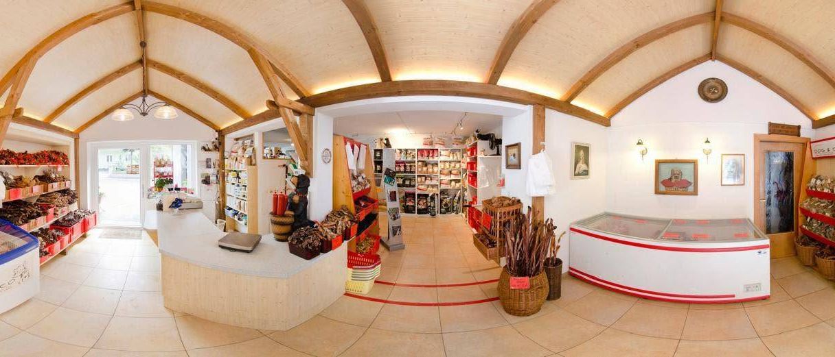 Ein Panoramablick auf einen Innenladen mit Holzdecke und gefliestem Boden. Eine Theke mit einer Waage und Körben ist auf der linken Seite. Regale mit Produkten und einem Ventilator befinden sich in der Mitte. Rechts an der Wand steht ein Kühlschrank und Bilderrahmen.