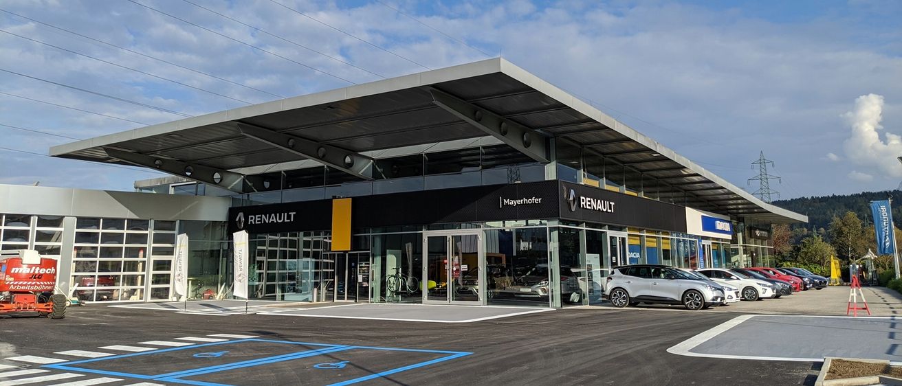 Das Äußere eines Renault-Händlers mit modernem Design, ausgestattet mit Glaswänden, einem großen Vordach und geparkten Autos davor. Der Himmel ist blau mit verstreuten Wolken.