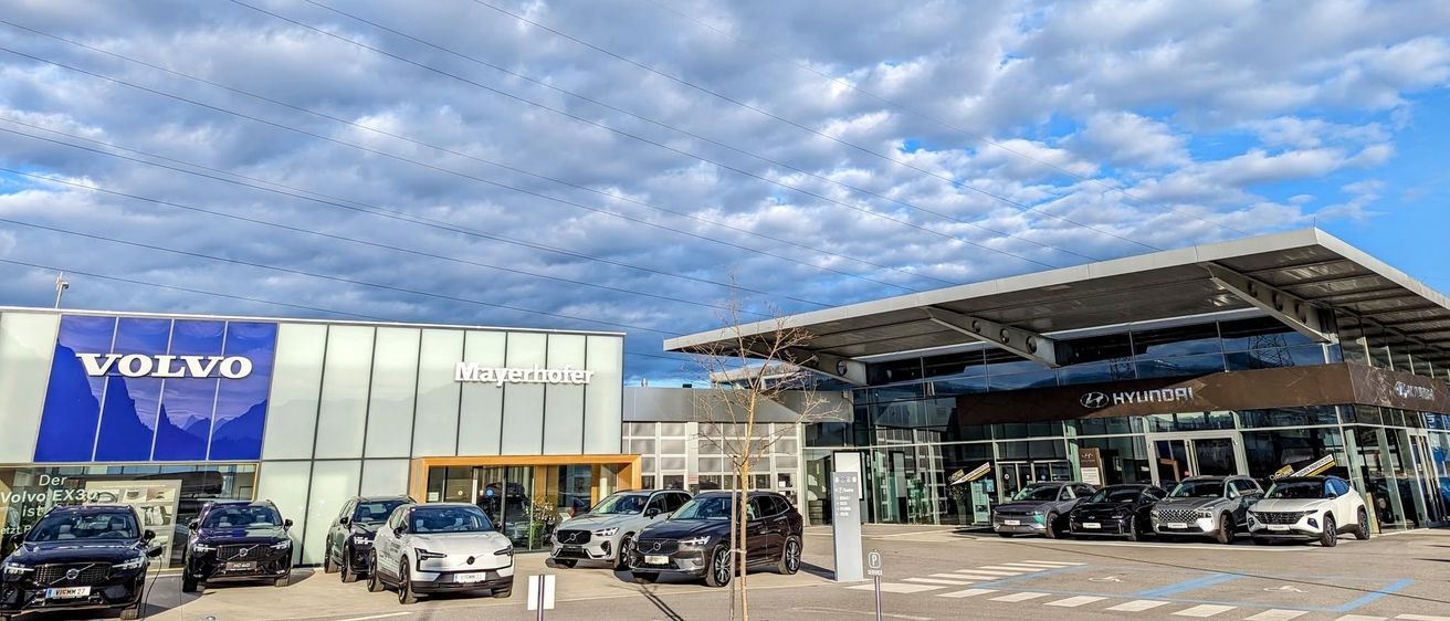 Ein modernes Autohaus mit mehreren Autos, die vor dem Gebäude unter einem bewölkten Himmel geparkt sind. Das Gebäude hat eine Glasfassade und ein Schild mit der Aufschrift 'Mayerhofer'.