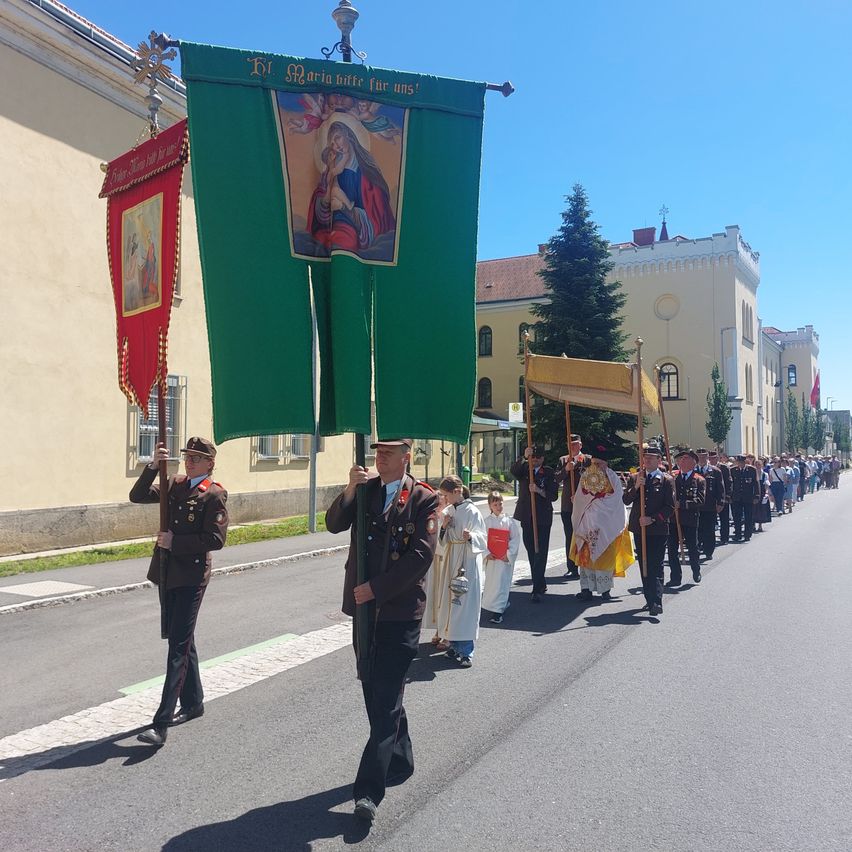 A procession of clergy and military officials walk in a parade with banners in front of a church and buildings.