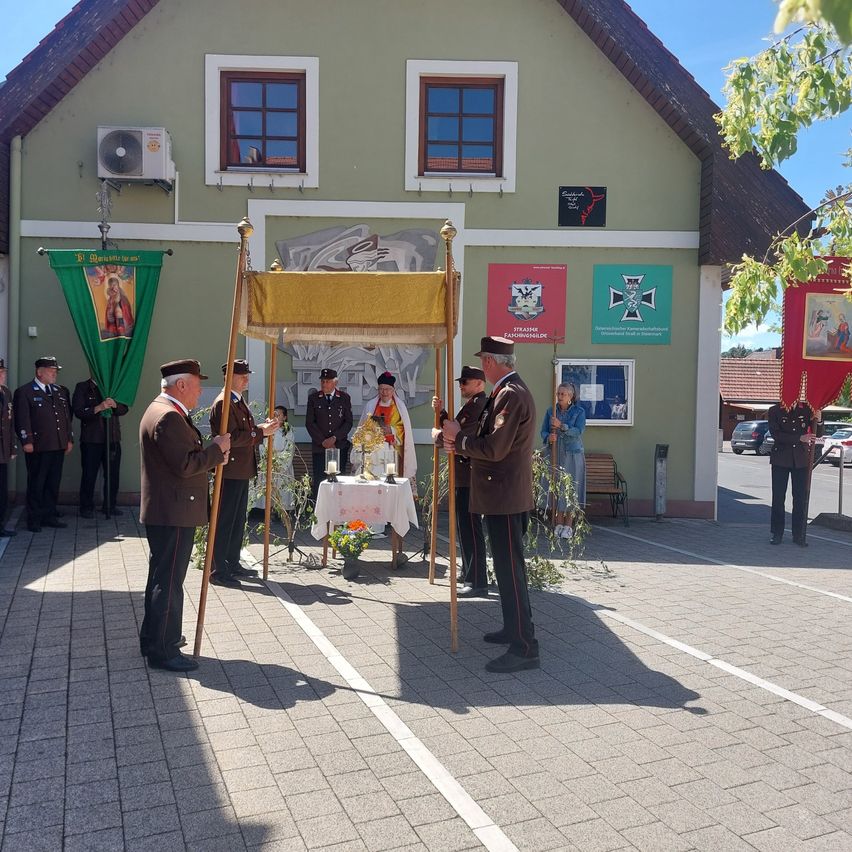 Uniformed men stand around a table in a ceremony, with an altar in the middle and flags on the side. Behind them is a building with banners and windows.