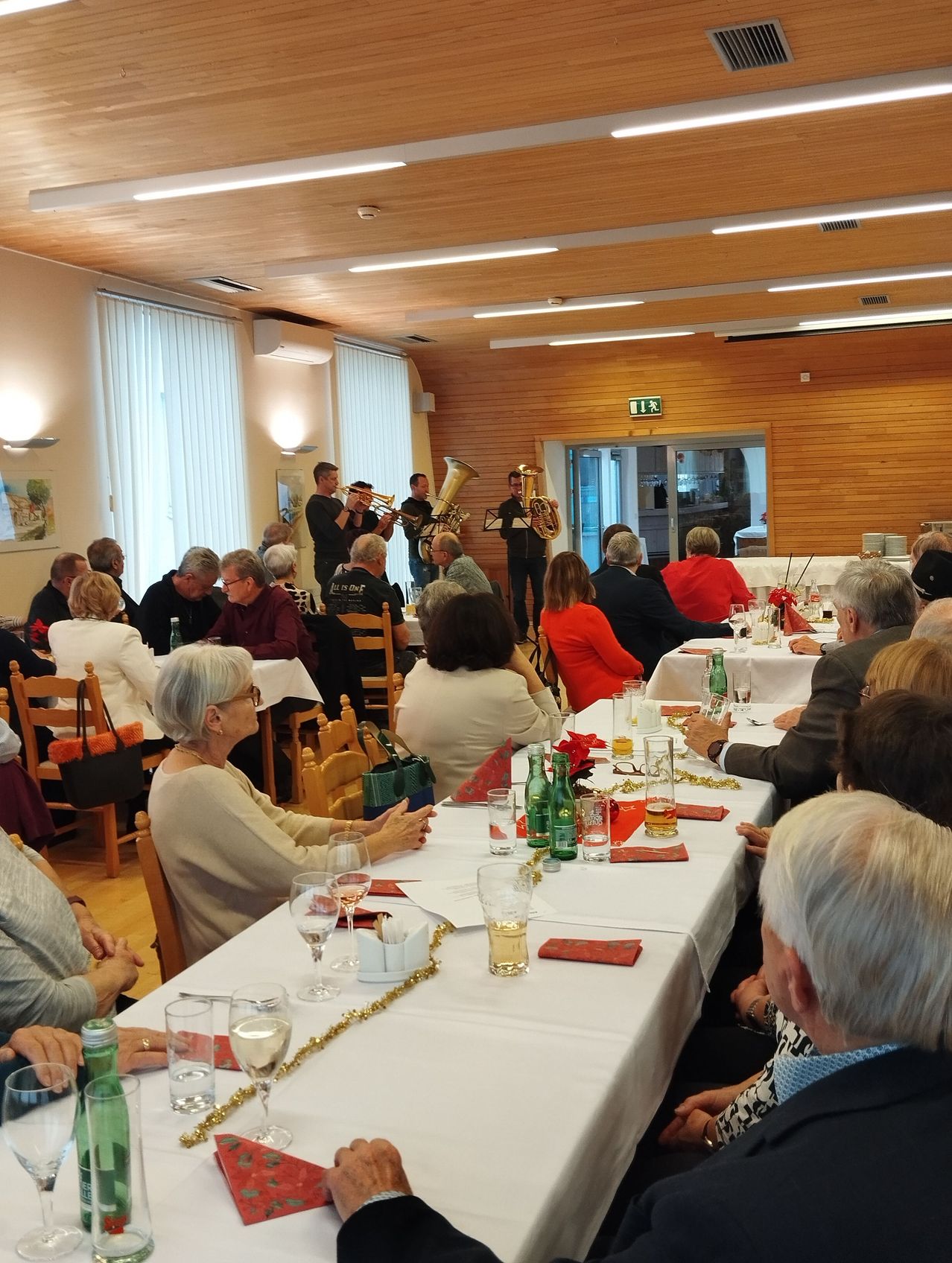 Eine Gruppe von Menschen versammelt sich in einem Speisesaal für eine formelle Veranstaltung. Einige sitzen, andere stehen. Eine Band spielt im Hintergrund auf Blechblasinstrumenten.