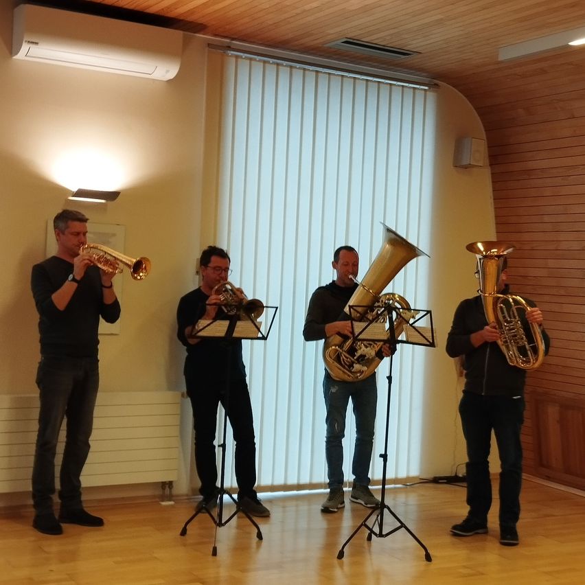 Vier Männer stehen in einem Raum und spielen Blechblasinstrumente. Sie stehen vor einem großen Fenster mit weißen Jalousien. Dahinter befindet sich eine Holzwand und eine an der Decke montierte Lampe.