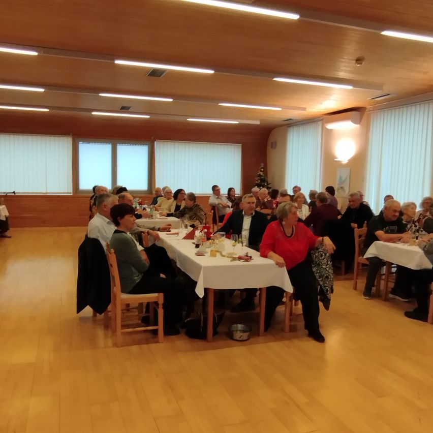 Eine Gruppe von Menschen sitzt an Tischen in einem Speisesaal. Die Tische sind mit weißen Tüchern bedeckt. Der Raum hat Holzböden und weiße Jalousien an den Fenstern. Eine Weihnachtsbaum steht in der Ecke.