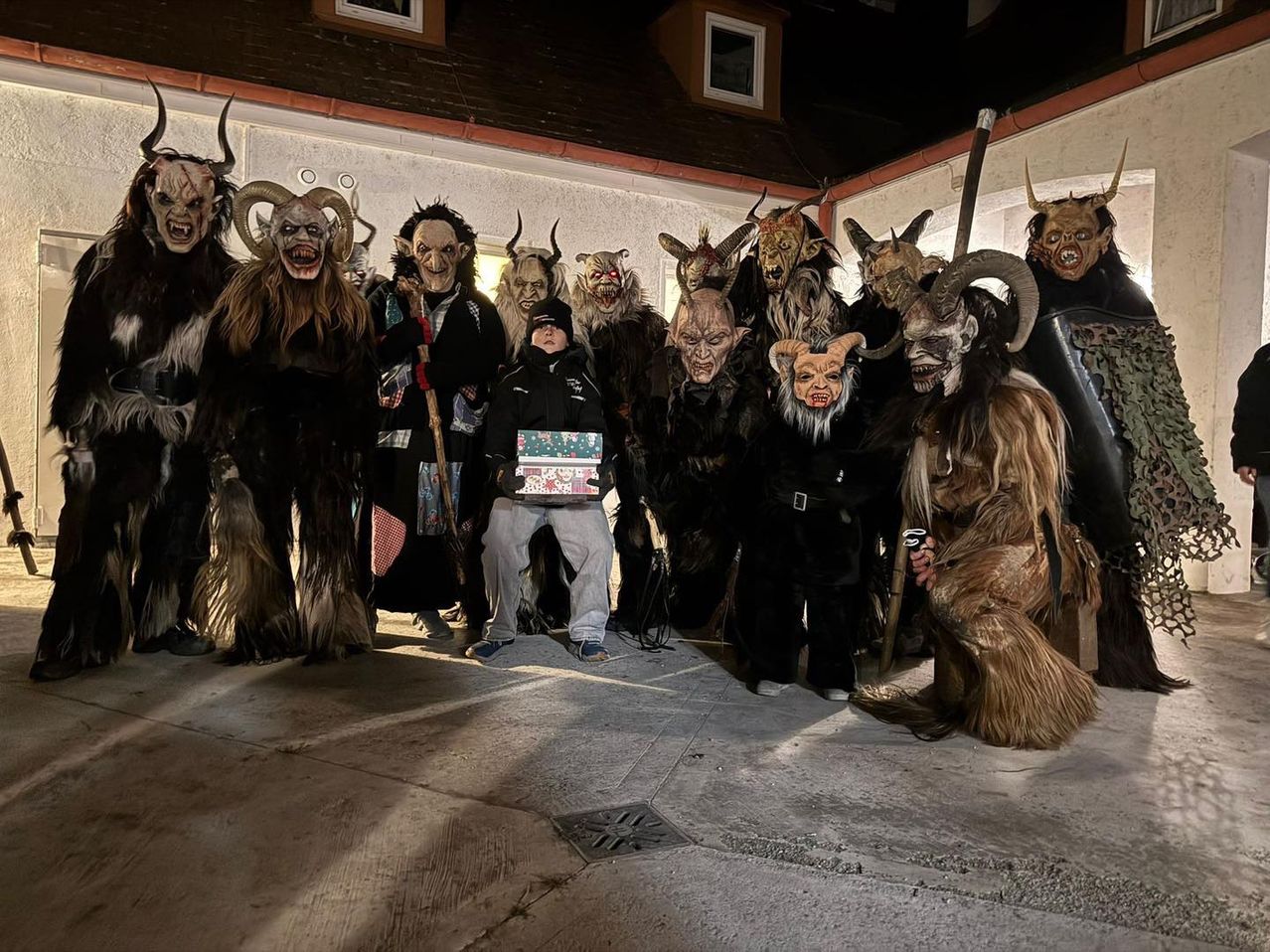 Eine Gruppe von Menschen in Hörnerkostümen steht mit einer Person, die einen Geschenkkarton hält, bei Nacht beleuchtet.