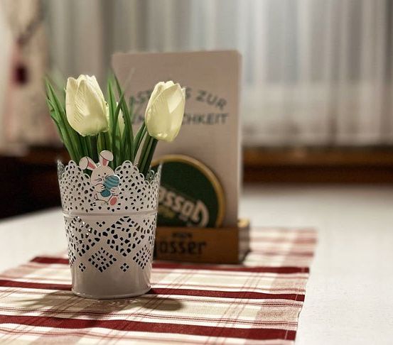 Ein Bouquet aus weißen Tulpen in einem dekorativen Glas auf einem Tisch mit rot-weiß kariertem Tuch.