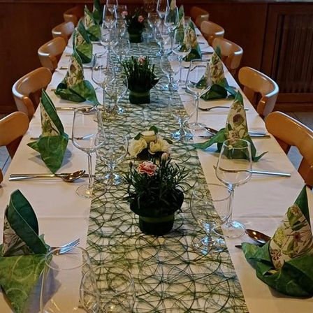 Ein langer Esstisch ist mit grünen Servietten, Weingläsern, Löffeln und Gabeln gedeckt. Ein Blumen-Mittelstück ist zwischen den Gläsern platziert.