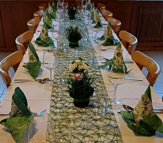 Ein langer Esstisch ist mit grünen Servietten, Weingläsern, Löffeln und Gabeln gedeckt. Ein Blumen-Mittelstück ist zwischen den Gläsern platziert.
