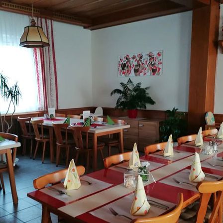 Ein gut eingerichtetes Restaurant mit einem langen Tisch, Stühlen und dekorativen Servietten. Der Raum ist gut beleuchtet, mit Pflanzen und einem Gemälde an der Wand.