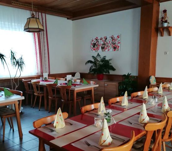 Ein gut eingerichtetes Restaurant mit einem langen Tisch, Stühlen und dekorativen Servietten. Der Raum ist gut beleuchtet, mit Pflanzen und einem Gemälde an der Wand.