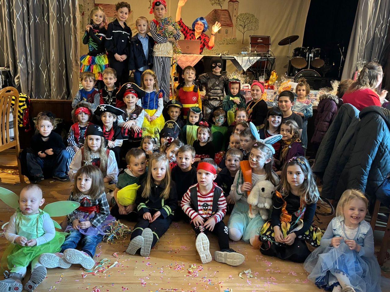 A group of children in various costumes, some standing and some sitting, are posing for a photo in a room with musical instruments in the background.