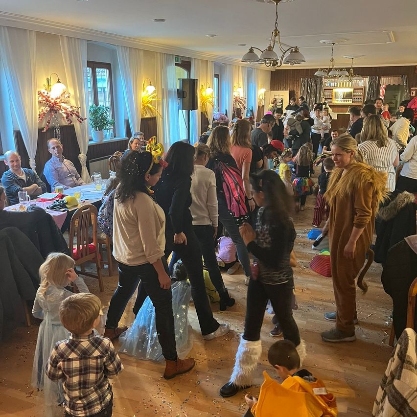 An indoor event with adults and children dancing, some dressed in costumes. A table with drinks and food is set up with chairs. There are lamps and plants in the room.