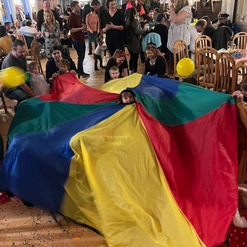 A group of adults and children play with a colorful parachute indoors. Some are peeking out from underneath it while others hold balloons.