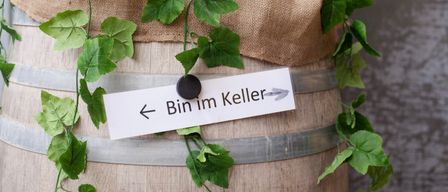 Ein mit Sackleinen bedeckter Fass mit grünen Blättern steht auf einer Holzoberfläche. Ein weißes Papier mit einem Pfeil und Text ist am Fass befestigt.