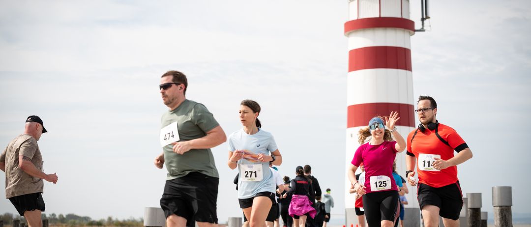 Eine Gruppe von Läufern mit Nummern nehmen an einem Rennen auf einem Pier teil, mit einem Leuchtturm im Hintergrund.