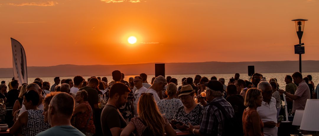 Eine große Menschenmenge versammelt sich bei Sonnenuntergang am Strand, mit Blick auf den Ozean und Berge in der Ferne. Der Himmel ist orange und rosa mit einer untergehenden Sonne.