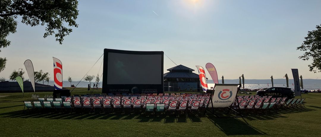 Eine große Outdoor-Kinoaufstellung mit Stuhlreihen unter klarem Himmel. Eine Projektionsleinwand ist sichtbar, und es gibt Fahnen und Banner mit Logos im Hintergrund.